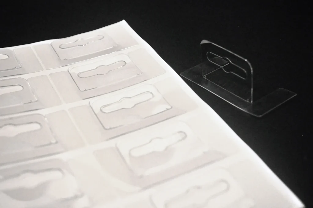 Clear PET self-adhesive Euro hole hang tabs on sheet, with a folded sample showing the hook slot design for retail packaging display.
